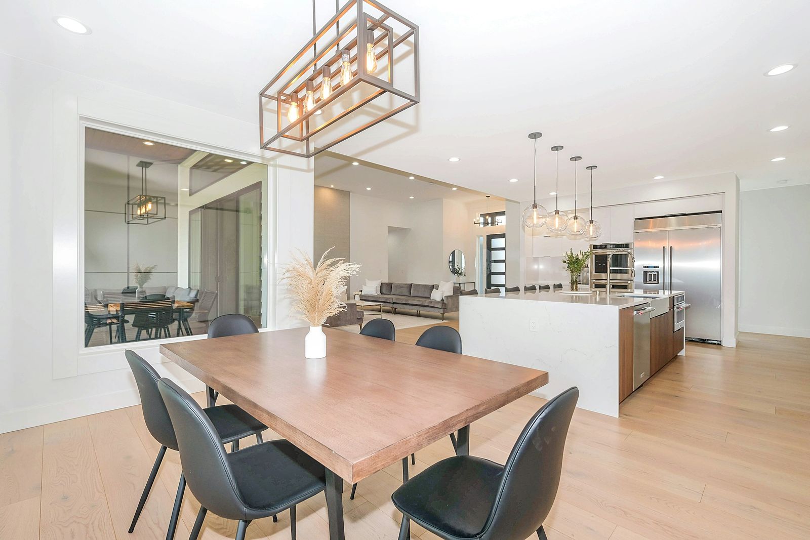 Dining area with a wood table under a rectangular cage linear chandelier, open to a kitchen with glass pendants and recessed lights