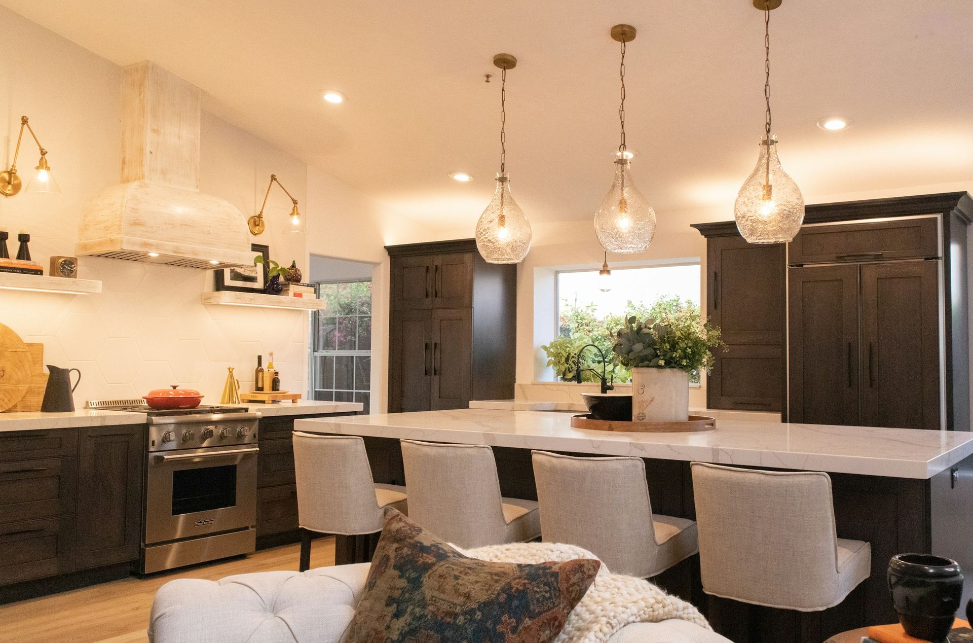 Warm evening kitchen with three glowing seeded-glass pendants over a white quartz island, brass swing-arm wall sconces, recessed downlights and LED strips under floating shelves