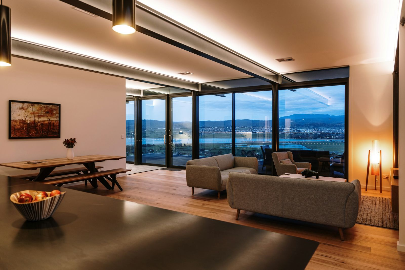 Open living and dining room at dusk with warm LED cove lighting along the ceiling beams, cylinder pendants and floor-to-ceiling windows