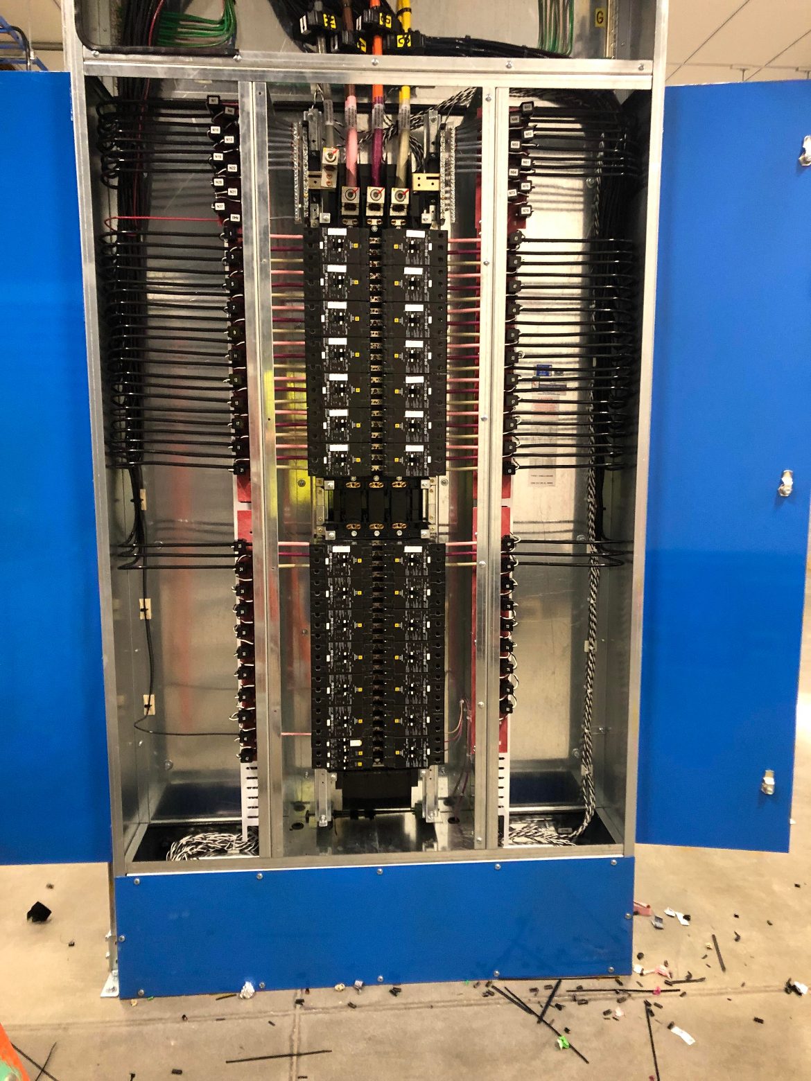 Tall commercial panelboard with blue doors open, rows of breakers down the center and branch wiring neatly bundled along both gutters