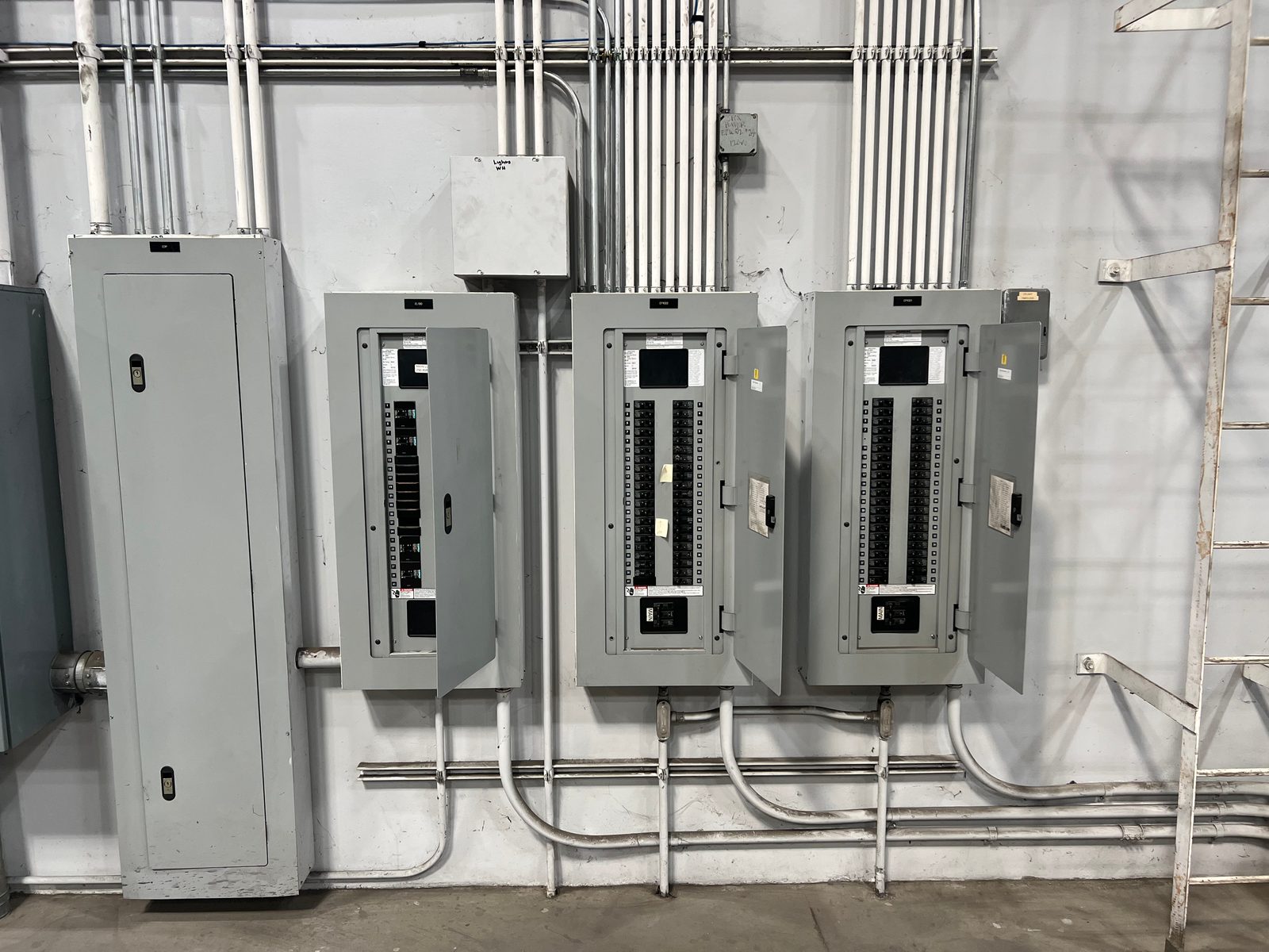 Three gray commercial panelboards with doors open and a tall enclosure, fed by parallel EMT conduit running along a block wall
