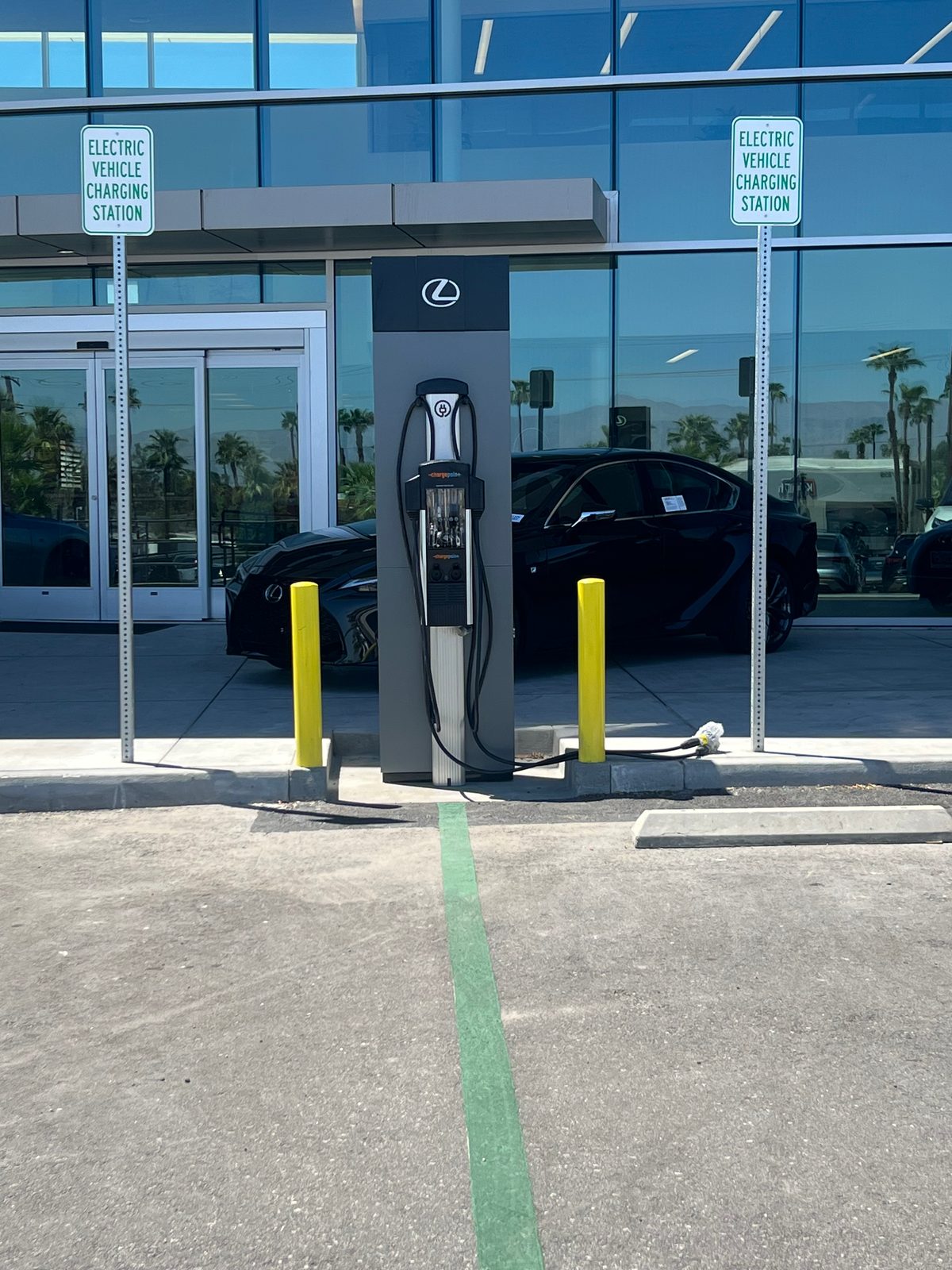 Pedestal EV charging station with yellow bollards and electric vehicle charging signs in front of a car dealership showroom