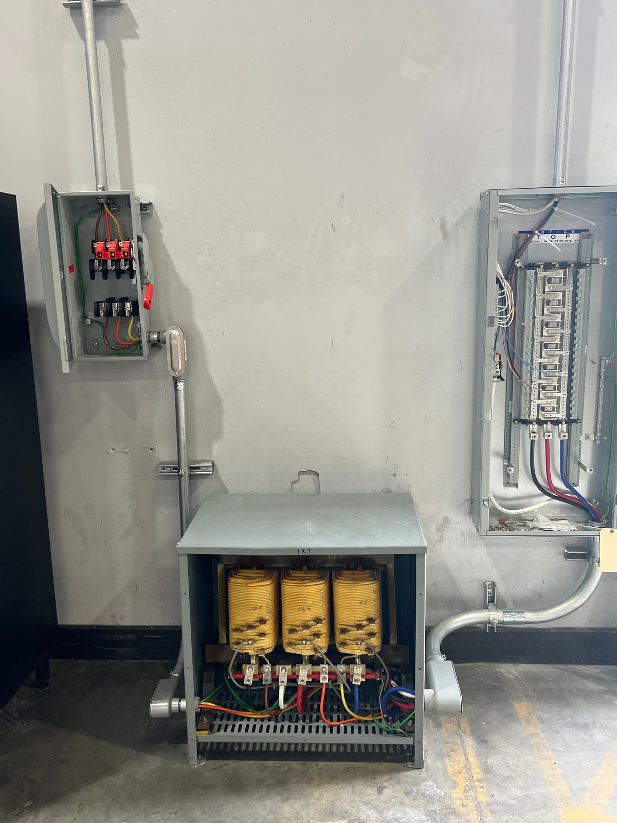 Dry-type transformer on the floor with a fused disconnect switch above left and an open panelboard on the right, connected with conduit
