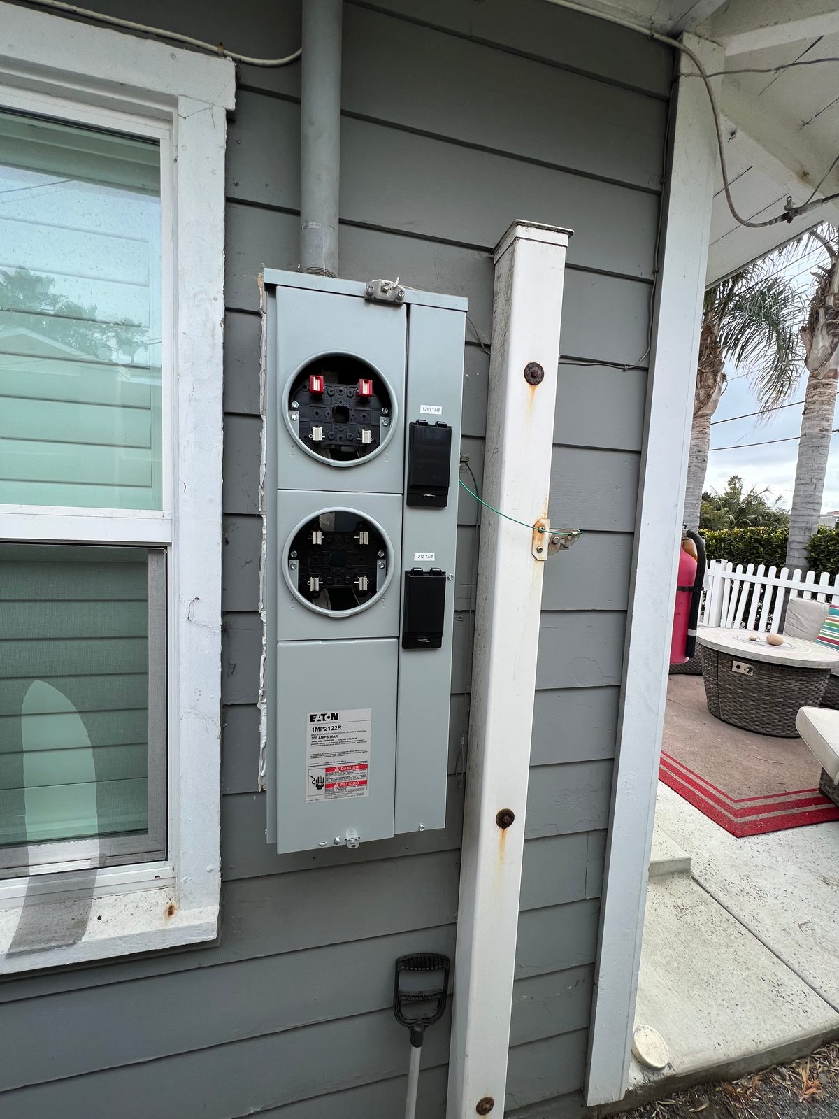 Gray two-position meter socket with blank covers mounted on the siding of a small house, beside a porch