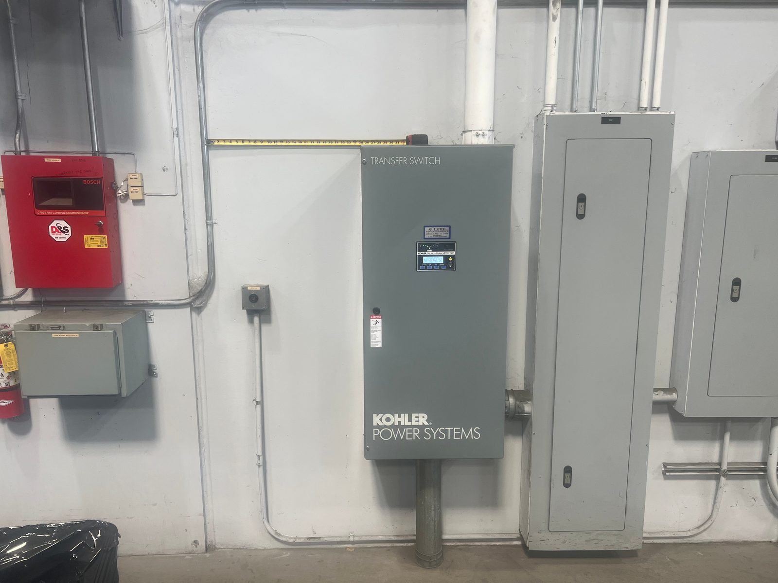 Gray Kohler automatic transfer switch mounted on a wall between a fire alarm panel and a distribution panel, all fed in EMT