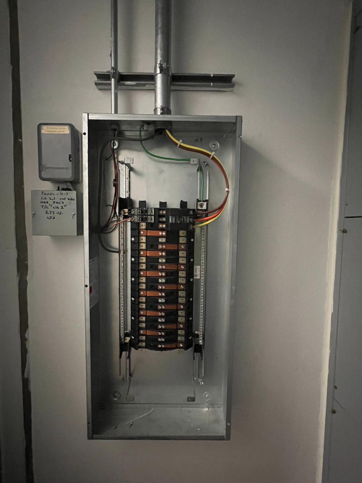 Gray panelboard with the cover off showing two rows of breakers, fed by conduit from the ceiling, with a small enclosure beside it