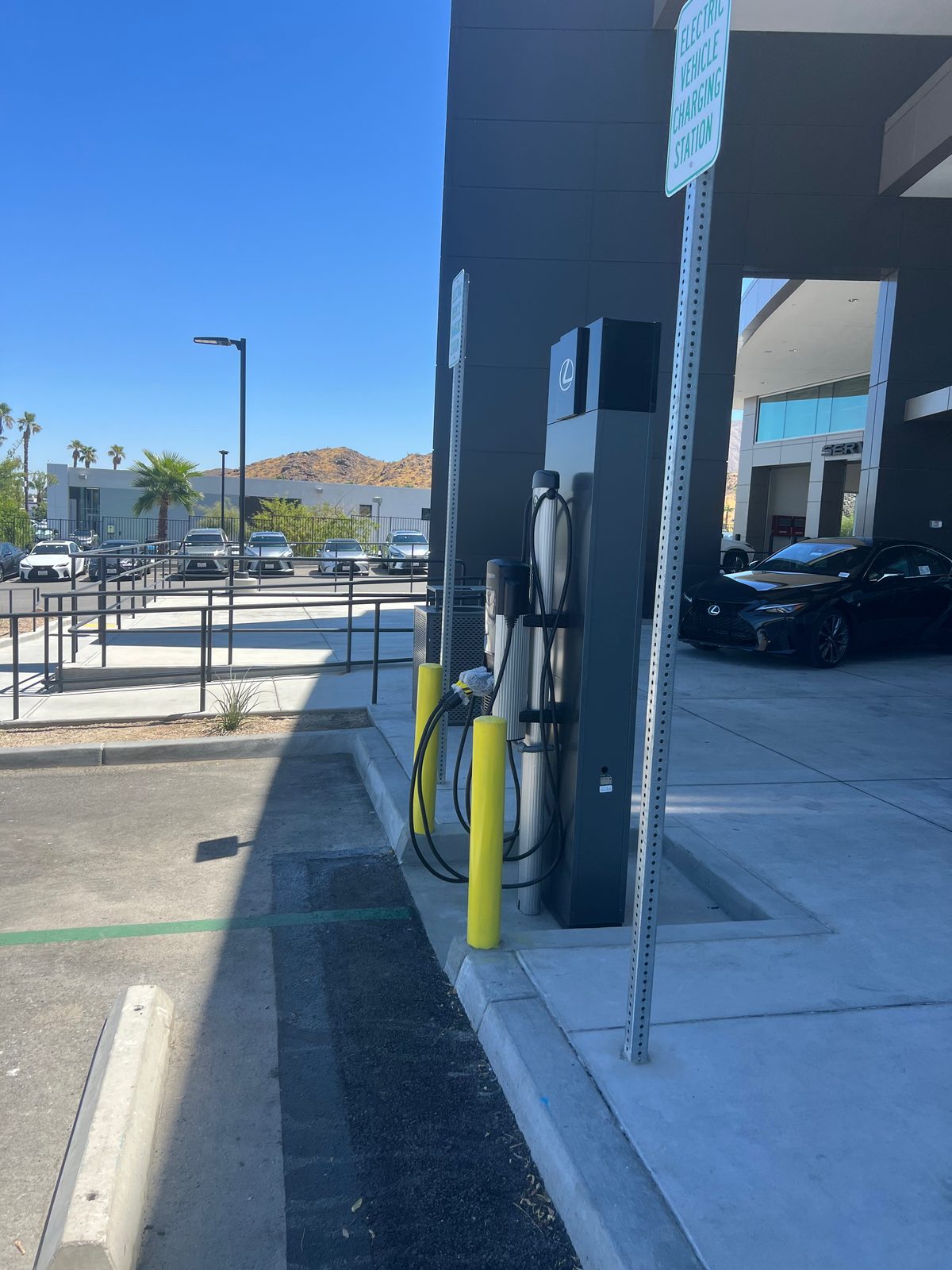 Dark pedestal EV charger with its cable holstered, protected by yellow bollards on a sidewalk beside a parking stall