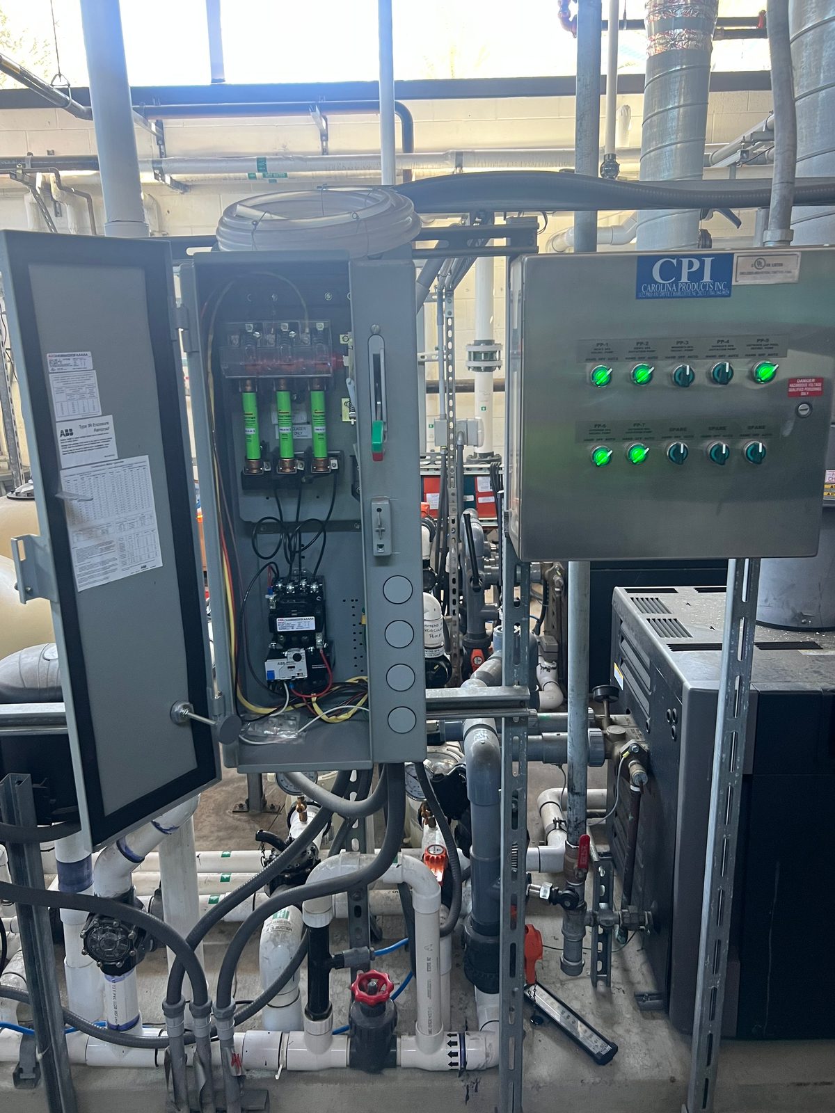 Open motor starter enclosure beside a stainless control panel with green indicator lights, above commercial pool pumps and plumbing