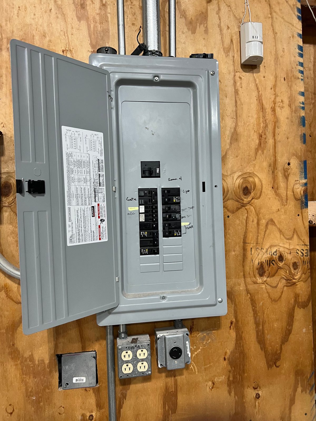 Gray load center with its door open on a plywood backboard, with duplex receptacles and a switch box mounted below