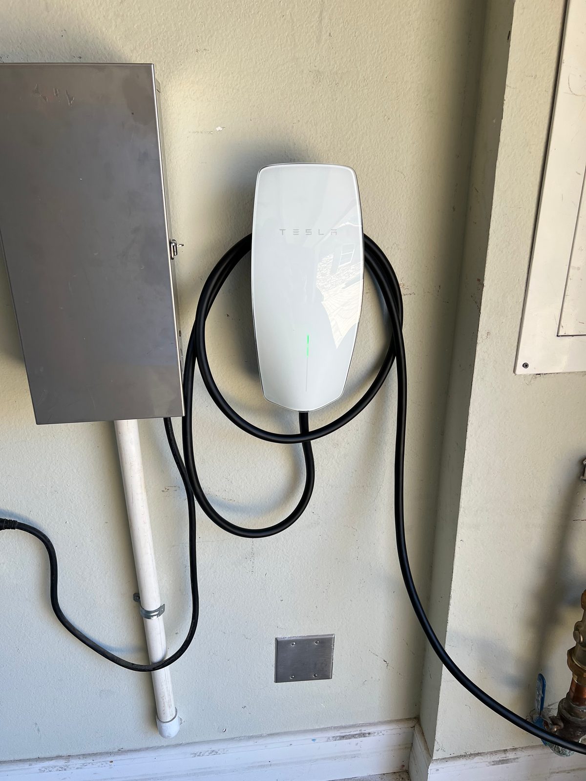 Tesla Wall Connector with a coiled cable mounted beside a gray panel cover, with conduit running up the wall