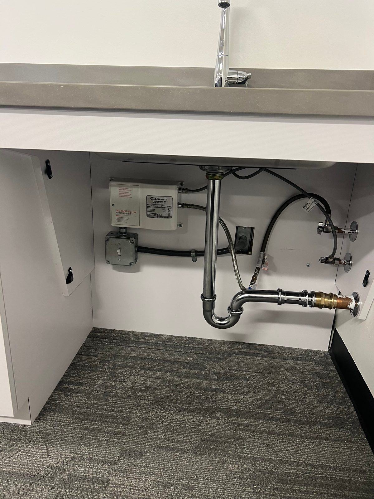 Under a sink: a point-of-use water heater mounted on the cabinet wall with a junction box and conduit feeding it, beside the plumbing