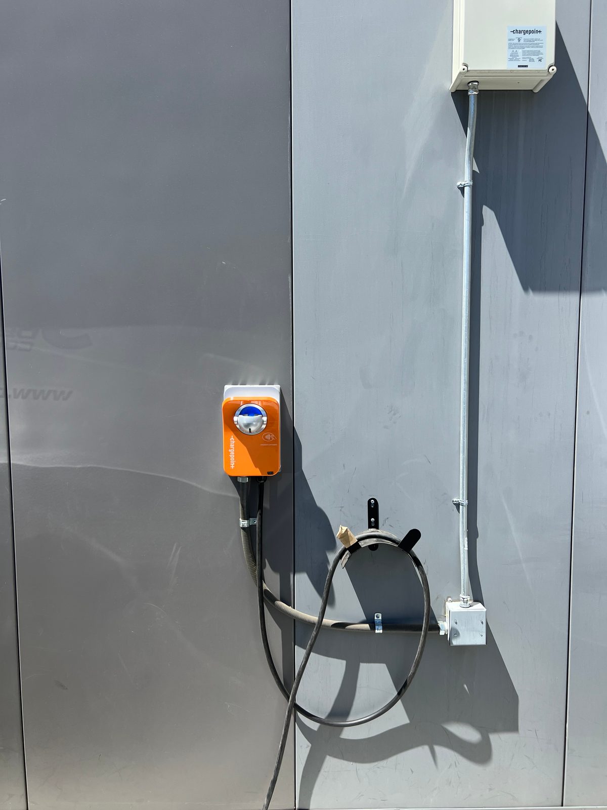 Orange wall-mounted EV charger on a gray building with its cable holstered, fed by conduit from a disconnect box above