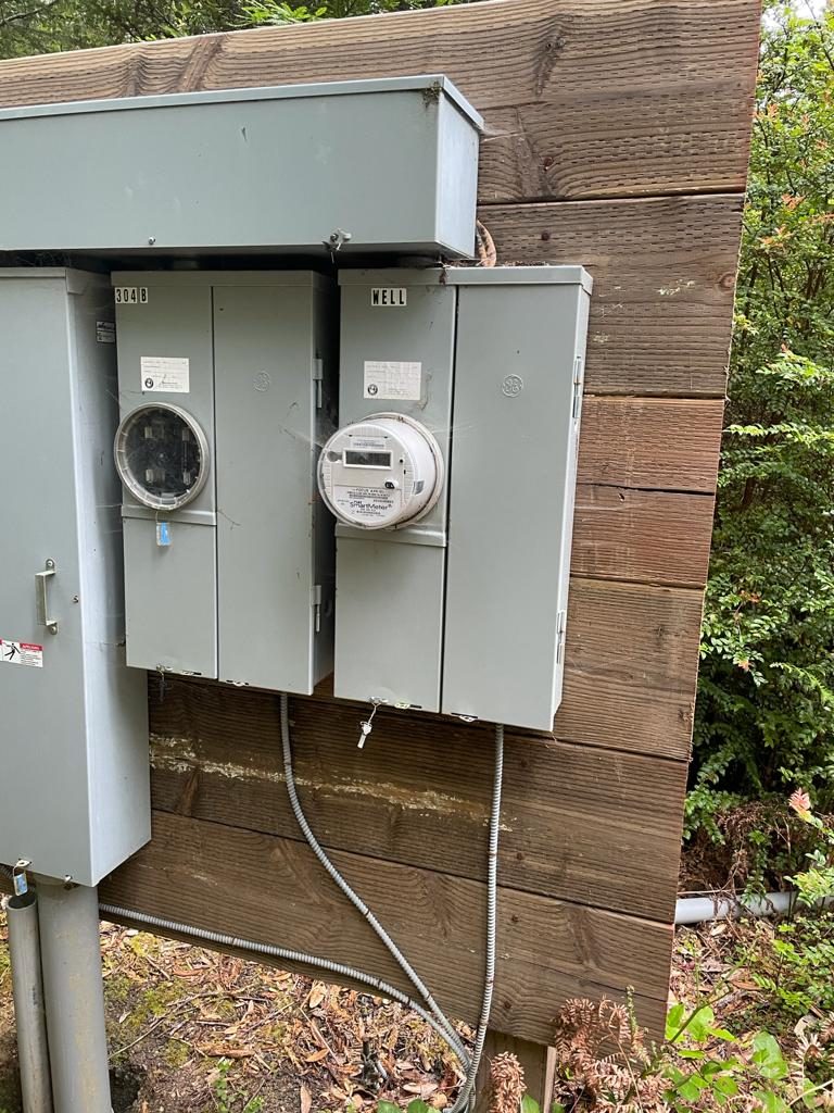 Two gray meter enclosures on a redwood-plank backboard under a wireway, one labeled 304B and one labeled WELL, with flexible conduit below
